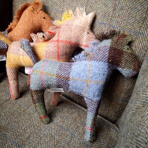 May include: Three plush horse toys made of tweed fabric in brown, beige, and blue plaid patterns. The horses are sitting on a tweed fabric surface.
