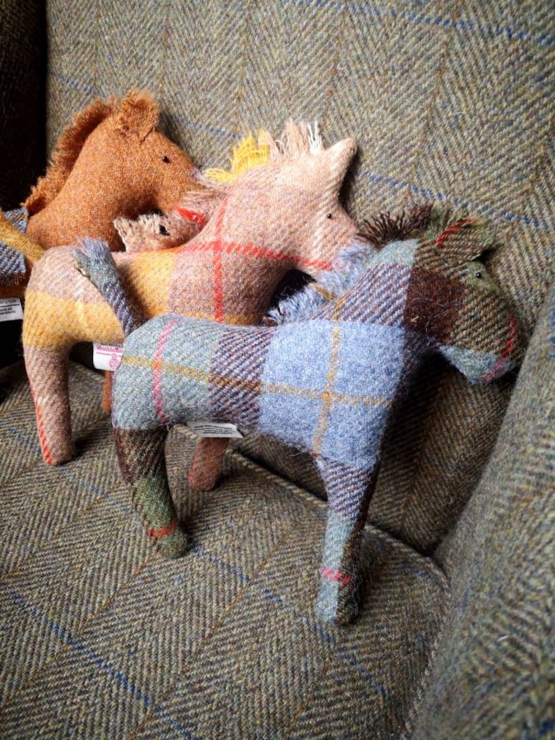 May include: Three plush horse toys made of tweed fabric in brown, beige, and blue plaid patterns. The horses are sitting on a tweed fabric surface.