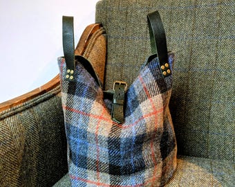 May include: A plaid bucket bag in shades of blue, gray, and red, with black leather handles and a buckle closure. The bag is made of a textured fabric and sits on a tweed armchair.