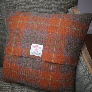 Puede incluir: Cojín de tiro de cuadros naranja, marrón y verde con una etiqueta "Harris Tweed". La etiqueta es blanca con texto negro y un pequeño logotipo.