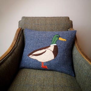May include: A blue and green patterned throw pillow with an embroidered image of a mallard duck. The pillow is sitting on a brown tweed armchair.