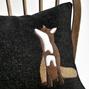Puede incluir: Una almohada negra con una aplicación de zorro marrón y blanco. La almohada está sentada en una silla de madera.