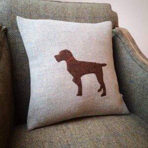 May include: A light blue throw pillow with a brown dog silhouette embroidered on it. The pillow is sitting on a green tweed armchair.