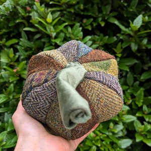 May include: A patchwork pumpkin made from tweed fabrics in brown, green, and blue hues. The pumpkin features a green fabric stem. The object is held in a hand against a backdrop of green foliage.