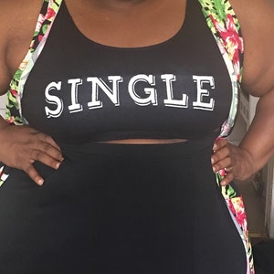 May include: A person wearing a black tank top with the word "SINGLE" printed on it. They are also wearing a floral print cardigan and a black skirt.