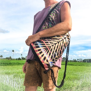 May include: A person wearing a purple shirt with a colorful tropical print and a black drawstring bag with a similar print. The bag is worn over the shoulder and has a long black strap.