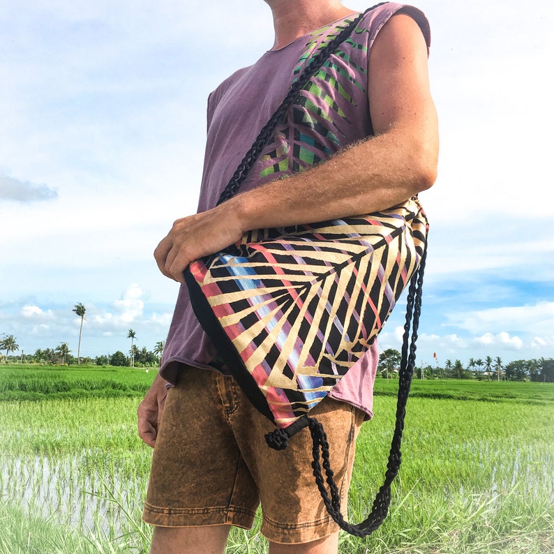 May include: A person wearing a purple shirt with a colorful tropical print and a black drawstring bag with a similar print. The bag is worn over the shoulder and has a long black strap.