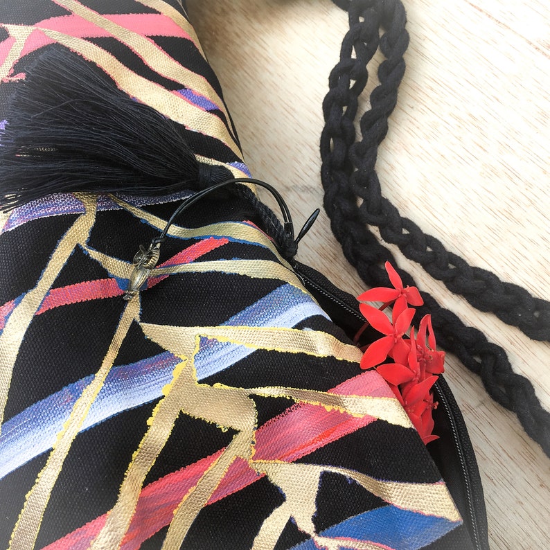 May include: A black canvas bag with a zipper closure and a black tassel. The bag has a geometric pattern of gold, red, and blue lines. The bag is made of a sturdy canvas material and has a long black strap.