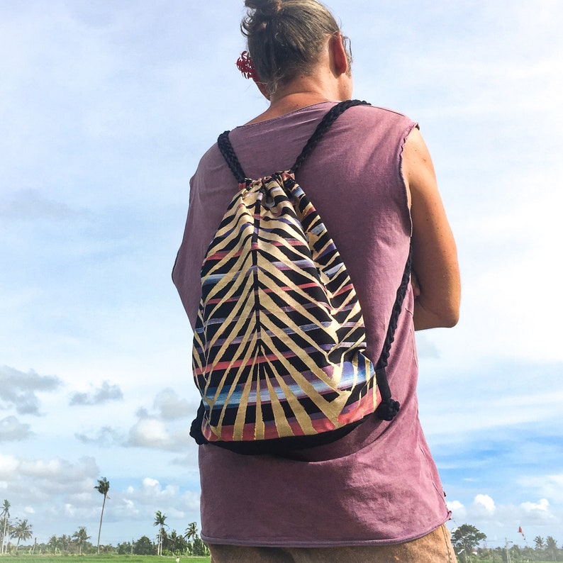 May include: A person wearing a purple tank top with a black drawstring bag with a colorful geometric pattern. The bag is worn on the back.