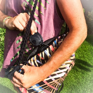 May include: A black and gold patterned tote bag with a black tassel. A person is holding the bag and a pair of black sunglasses are partially visible inside the bag.