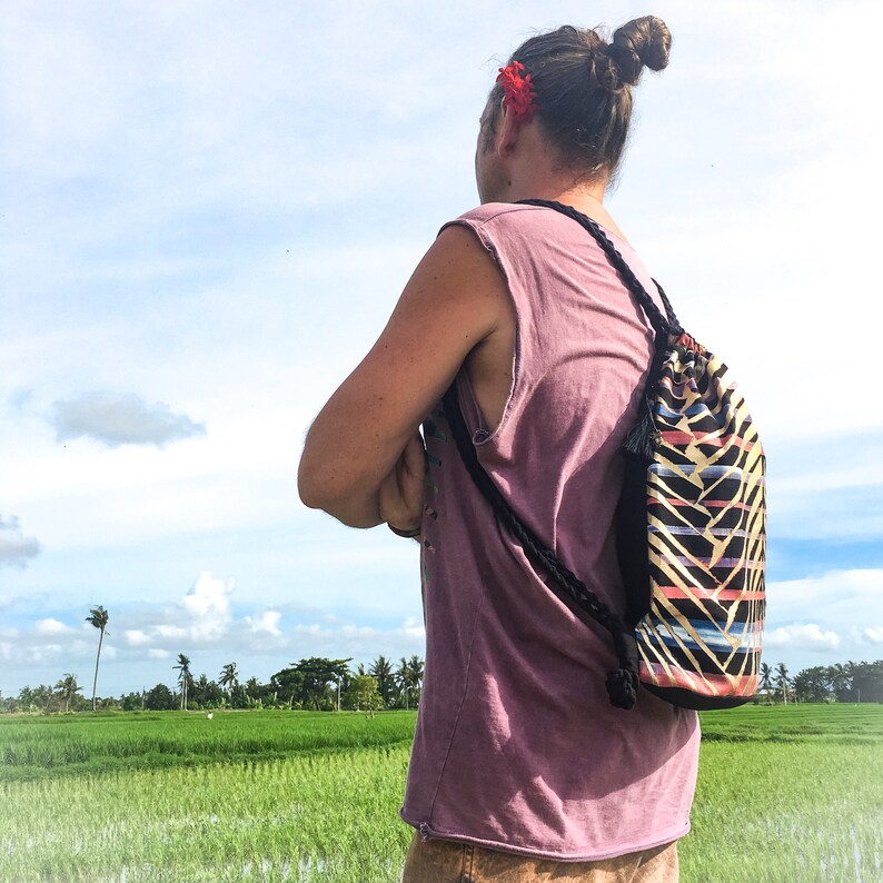 May include: A person wearing a pink tank top and a black drawstring bag with a colorful geometric pattern. The bag is worn on the back and has a long black strap. The person is standing in a field with green grass and a blue sky in the background.