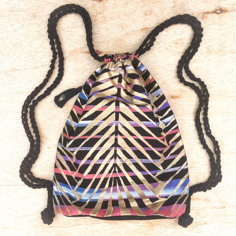 May include: A black drawstring bag with a geometric pattern in gold, pink, and blue. The bag has black drawstrings and a black tassel.