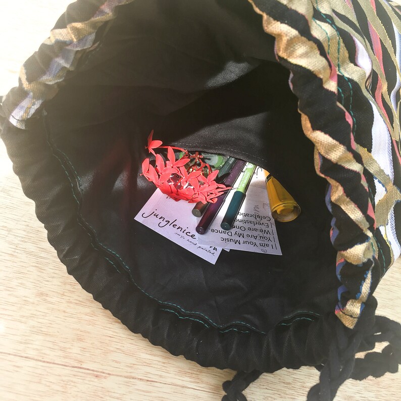 May include: A black drawstring bag with a colorful patterned exterior. The bag is open and the inside is lined with black fabric. The bag contains a few pens, a small bottle of liquid, and a red flower. A white card with the text "JungleNice" is visible inside the bag.