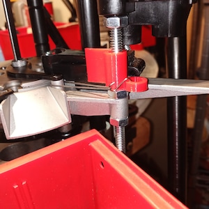May include: Close-up of a reloading press with a red plastic component and a silver metal screw. The press is mounted on a red plastic box.