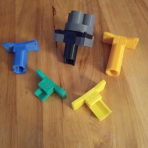 May include: A collection of 3D-printed plastic parts in various colors, including blue, green, yellow, and gray. The parts have a T-shape and rectangular shapes. Some parts have text printed on them. The objects are arranged on a wooden surface.