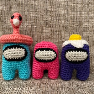 May include: Three crocheted Among Us characters in blue, pink, and purple. The blue character has a pink flamingo-like hat. The pink character is a simple crewmate. The purple character has a white and yellow flower hat. Each has a white oval visor.