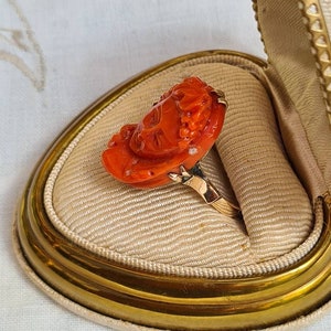 May include: A vintage ring featuring a carved orange coral rose design set on a gold band. The ring is displayed inside a cream-colored, heart-shaped jewelry box with a gold trim. The coral rose has intricate details, showcasing the craftsmanship of the piece.