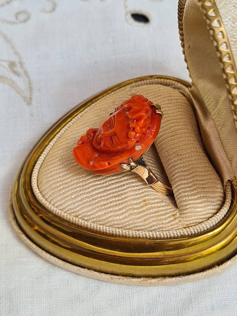 May include: A vintage ring featuring a carved orange coral rose design set on a gold band. The ring is displayed inside a cream-colored, heart-shaped jewelry box with a gold trim. The coral rose has intricate details, showcasing the craftsmanship of the piece.