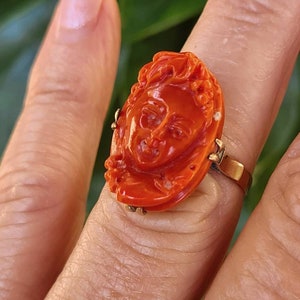 May include: A vibrant orange ring featuring a carved face design. The ring has a gold band and a detailed, oval-shaped face carving. The ring is a close-up shot, showcasing the intricate details of the face.