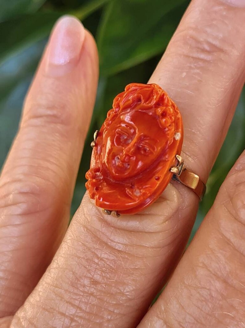 May include: A vibrant orange ring featuring a carved face design. The ring has a gold band and a detailed, oval-shaped face carving. The ring is a close-up shot, showcasing the intricate details of the face.