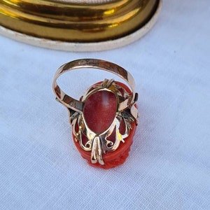 May include: A gold ring featuring a carved coral gemstone. The coral is a vibrant red-orange color, set in a detailed gold filigree setting. The ring's band is a simple, polished gold.