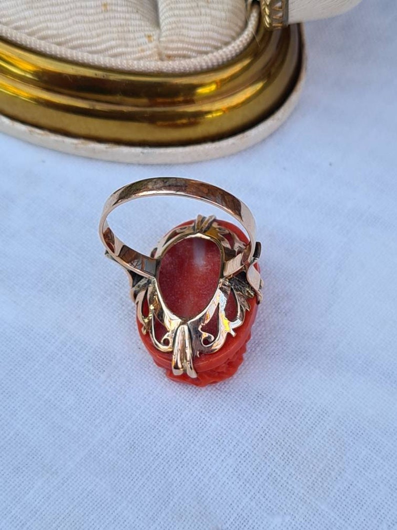 May include: A gold ring featuring a carved coral gemstone. The coral is a vibrant red-orange color, set in a detailed gold filigree setting. The ring's band is a simple, polished gold.