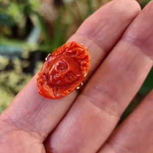 May include: A carved, orange-red ring with a detailed face design. The ring is set on a gold band. The ring's face is approximately 1 inch in length and 0.75 inches in width. The ring is held in an open hand.