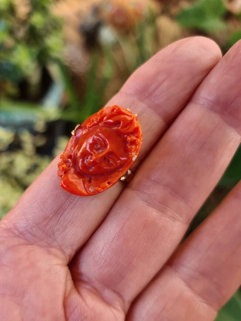 May include: A carved, orange-red ring with a detailed face design. The ring is set on a gold band. The ring's face is approximately 1 inch in length and 0.75 inches in width. The ring is held in an open hand.