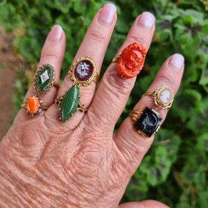 May include: A hand displaying a collection of antique rings. The rings feature various designs, including a carved face, a cameo, and gemstones in colors like green, orange, and black. The rings are set in gold-toned metal.