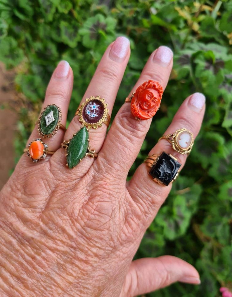 May include: A hand displaying a collection of antique rings. The rings feature various designs, including a carved face, a cameo, and gemstones in colors like green, orange, and black. The rings are set in gold-toned metal.