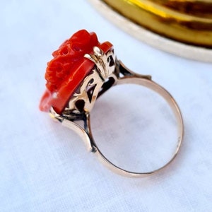 May include: A gold ring featuring a carved red coral rose. The ring has an ornate gold filigree setting and a delicate band. The rose is intricately detailed, showcasing the craftsmanship of the jewelry piece. The ring is set against a soft white background.