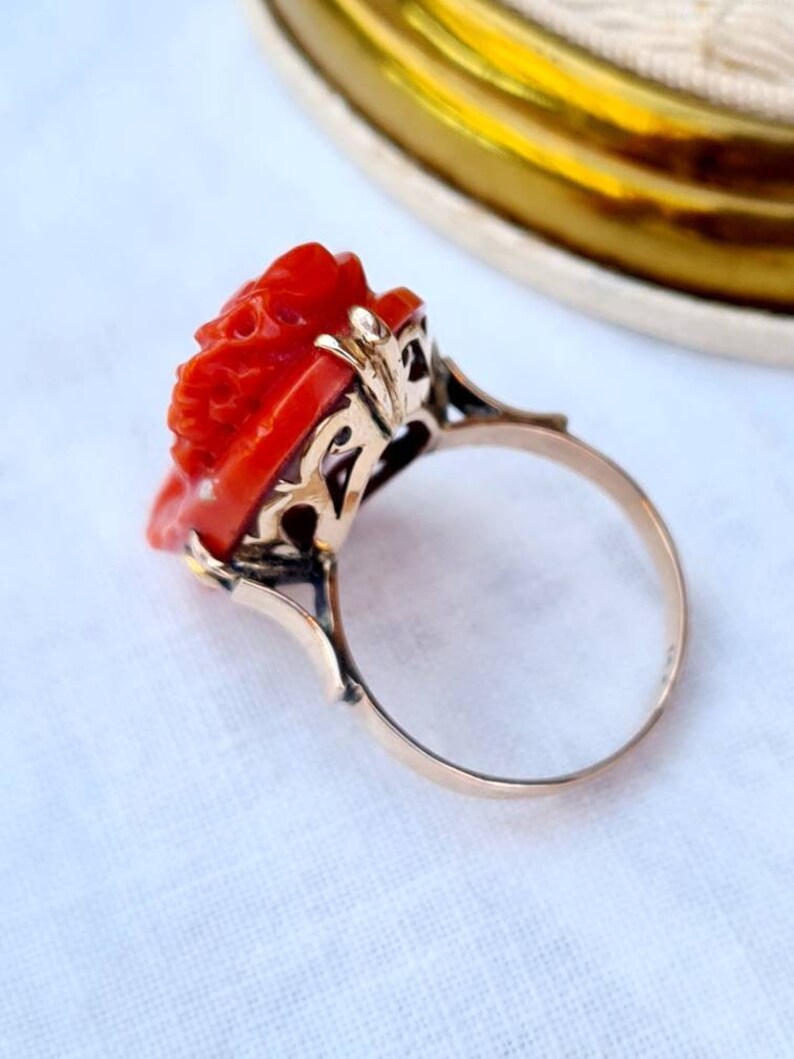 May include: A gold ring featuring a carved red coral rose. The ring has an ornate gold filigree setting and a delicate band. The rose is intricately detailed, showcasing the craftsmanship of the jewelry piece. The ring is set against a soft white background.