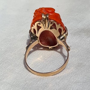 May include: A gold ring featuring a carved, vibrant orange gemstone. The gemstone is intricately sculpted with a floral or organic design. The ring's band and setting are a warm gold color, creating a striking contrast with the gemstone.
