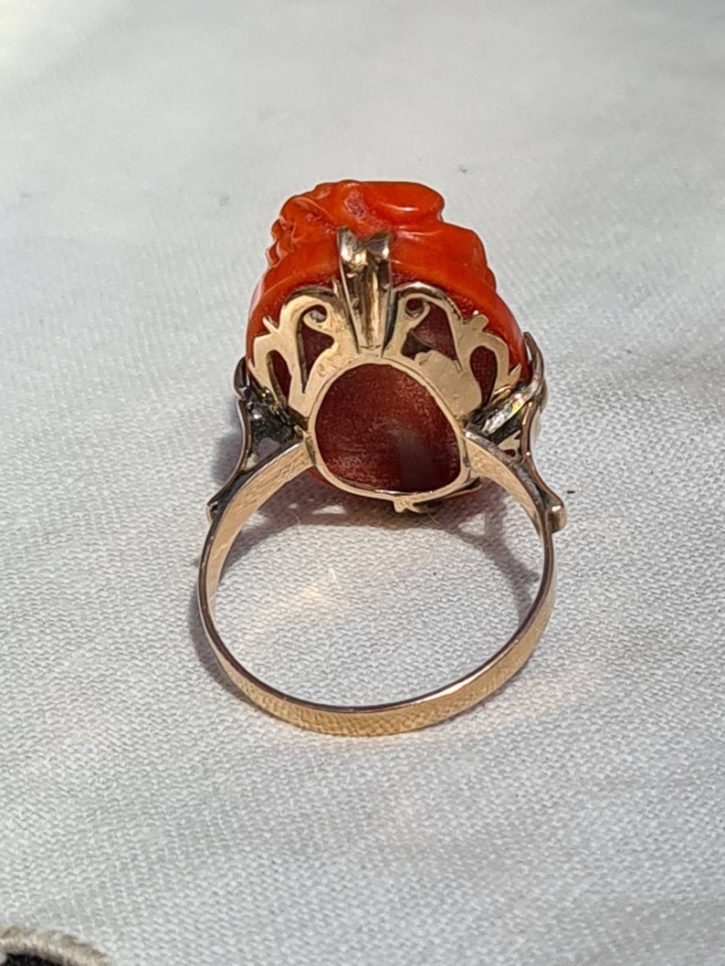 May include: A gold ring featuring a carved, vibrant orange gemstone. The gemstone is intricately sculpted with a floral or organic design. The ring's band and setting are a warm gold color, creating a striking contrast with the gemstone.