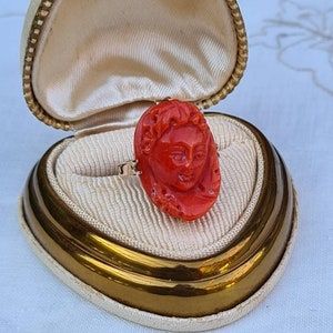 May include: A vintage ring featuring a carved red coral cameo of a classical face. The ring is set in gold and displayed in a cream-colored jewelry box with a gold trim and a cream satin interior. The box is open, showcasing the ring.