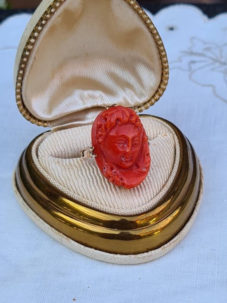 May include: A vintage ring featuring a carved red coral cameo of a classical face. The ring is set in gold and displayed in a cream-colored jewelry box with a gold trim and a cream satin interior. The box is open, showcasing the ring.