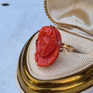 May include: A vintage gold ring featuring a carved coral cameo of a woman's face. The ring is set in a gold band and is displayed in a cream-colored jewelry box with a gold trim. The coral is a vibrant orange-red color.