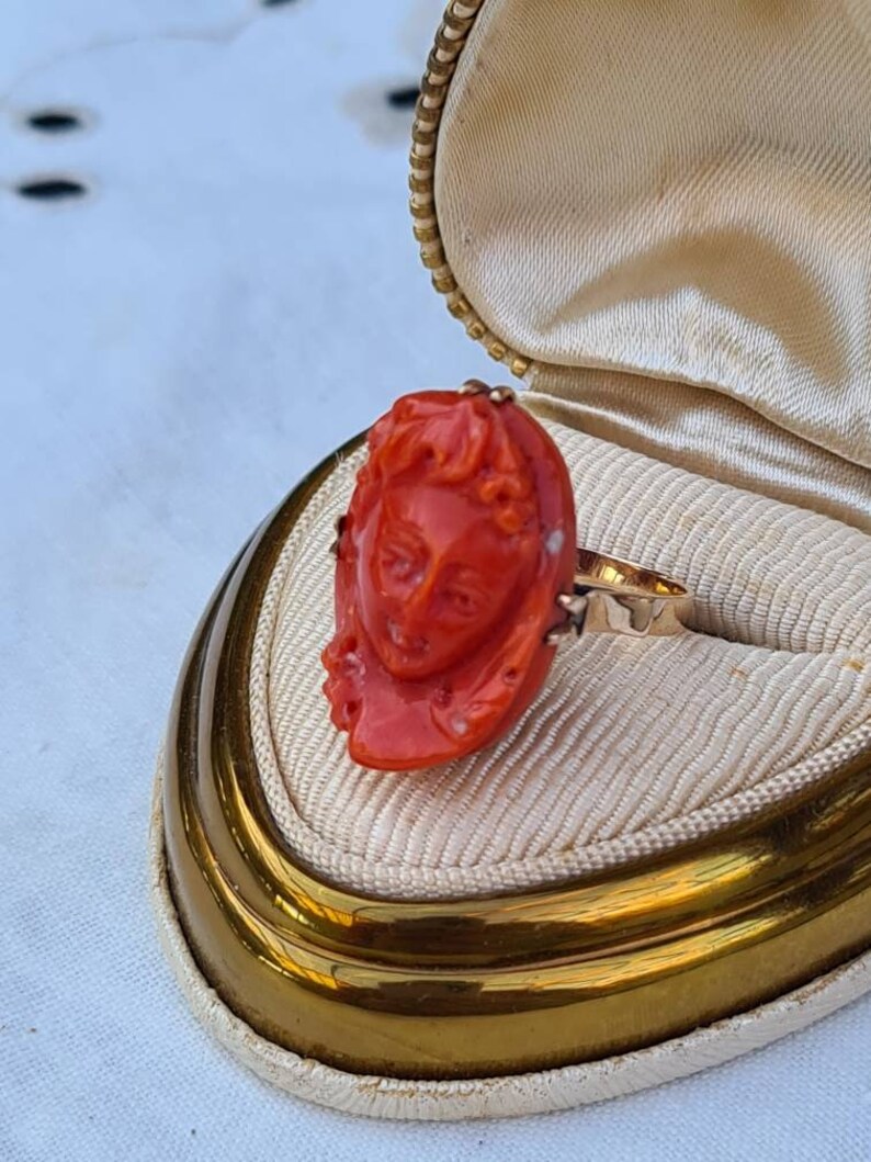 May include: A vintage gold ring featuring a carved coral cameo of a woman's face. The ring is set in a gold band and is displayed in a cream-colored jewelry box with a gold trim. The coral is a vibrant orange-red color.