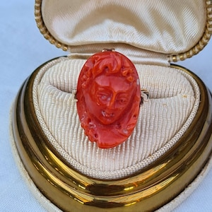May include: A vintage ring featuring a carved oval coral cameo of a woman's face. The ring is set in a gold band and displayed in a cream-colored jewelry box with a gold trim.