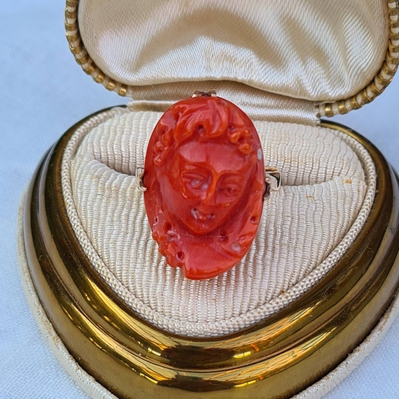 May include: A vintage ring featuring a carved oval coral cameo of a woman's face. The ring is set in a gold band and displayed in a cream-colored jewelry box with a gold trim.