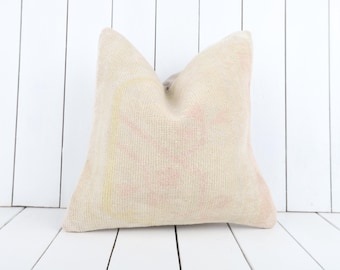 Puede incluir: Un cojín cuadrado de color crema con una superficie texturizada. El cojín presenta un patrón descolorido, similar a flores, en tonos rosa y amarillo. El cojín se coloca sobre un fondo de madera blanca.