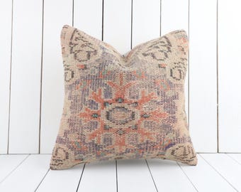 May include: Square cushion with a vintage design. The cushion has a central geometric pattern in shades of orange, purple, and beige. It is made of woven fabric and is set against a white wooden backdrop.