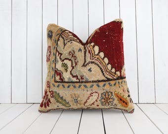 Puede incluir: Un cojín cuadrado con una funda de tejido textil con un patrón denso e intrincado. El diseño incorpora una sección de color rojo intenso que contrasta con un fondo beige, acentuado con motivos florales y de cachemira en azul, verde y naranja.