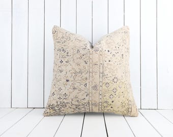 May include: A square beige pillow with a vintage-style pattern in shades of gray and cream. The pillow features a patchwork design with floral and geometric motifs, creating a textured appearance. The pillow is placed against a white wooden background.