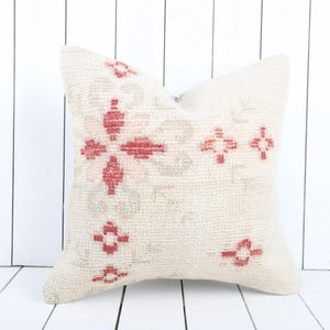 May include: Cream-colored decorative pillow with a geometric floral pattern in red. The pillow has a soft, textured appearance and is placed against a white wooden background. The design features a central flower and smaller cross-like shapes.