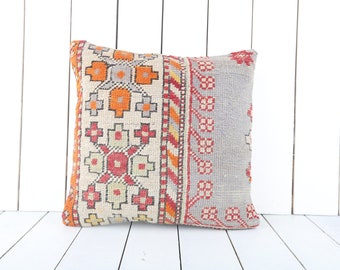 Puede incluir: Una almohada decorativa con fondo blanco y un patrón geométrico colorido en tonos rojos, naranjas y amarillos. La almohada está hecha de tejido tejido y tiene un borde con flecos.