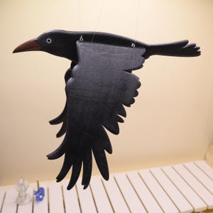 May include: A black wooden crow mobile with outstretched wings, suspended by thin strings. The crow has a brown beak and a detailed wing design, creating a decorative and artistic piece. The crow is a decorative item.