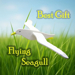May include: A white wooden seagull with yellow beak and wing details, depicted in flight. The background features green grass and a blue sky, with the words "Best Gift" and "Flying Seagull" in yellow text.