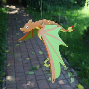 May include: A hanging wooden dragon mobile featuring an orange body with yellow spots and large, bright green wings with orange accents. The dragon has a long tail with a red tip and a small black flower-shaped detail hanging from its wing.
