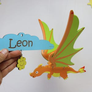 May include: A hanging mobile featuring an orange dragon with bright green wings and a light blue cloud-shaped wooden plaque with the name Leon engraved in brown. A small yellow flower-shaped charm with a smiling face hangs from the cloud.